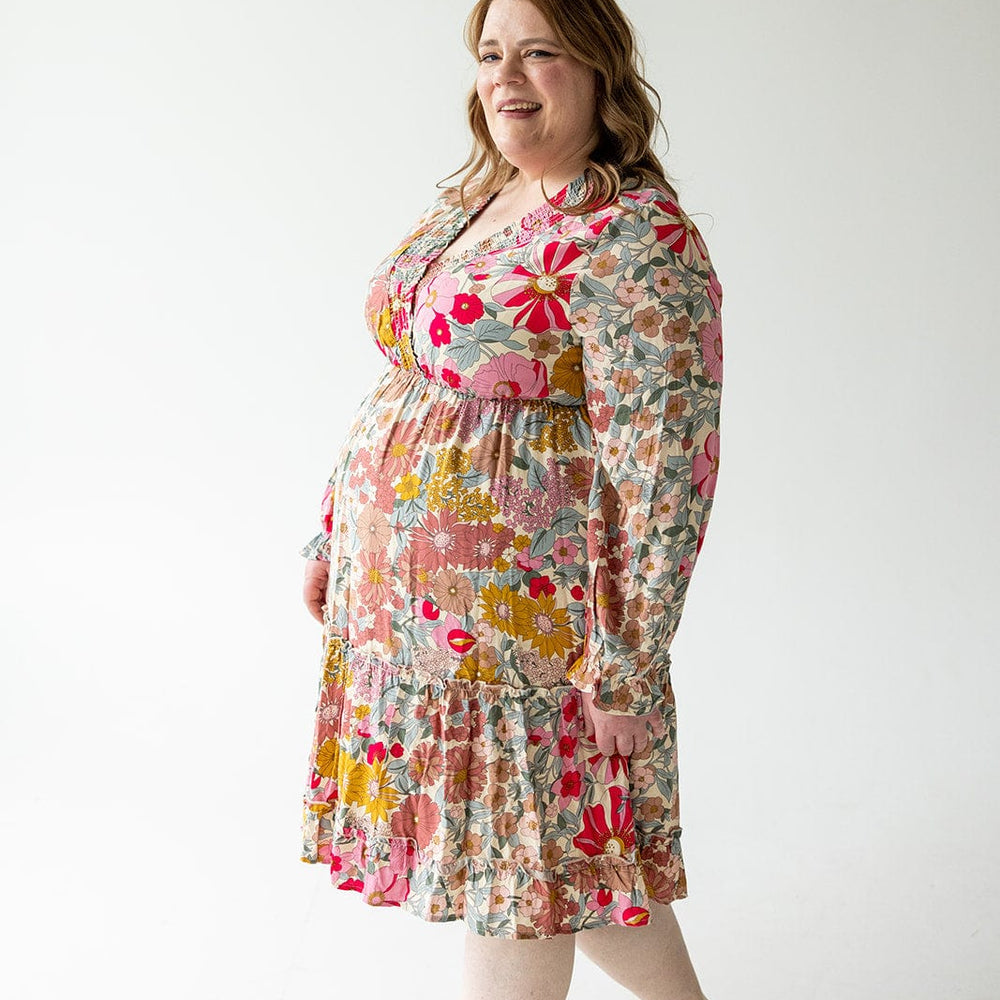 A woman in a SMOCKED BODICE BOHO DRESS by Love Marlow, which showcases a floral pattern, stands in a bright room with a white background, smiling and looking to the side.