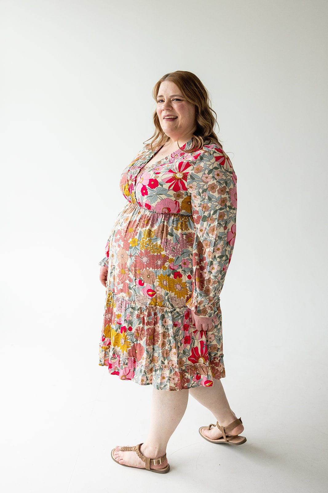 A woman in a SMOCKED BODICE BOHO DRESS by Love Marlow, which showcases a floral pattern, stands in a bright room with a white background, smiling and looking to the side.