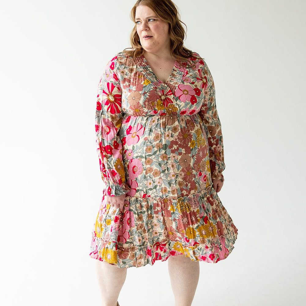 A person stands in Love Marlow's SMOCKED BODICE BOHO DRESS, showcasing pink, red, and yellow floral patterns, looking to the side.