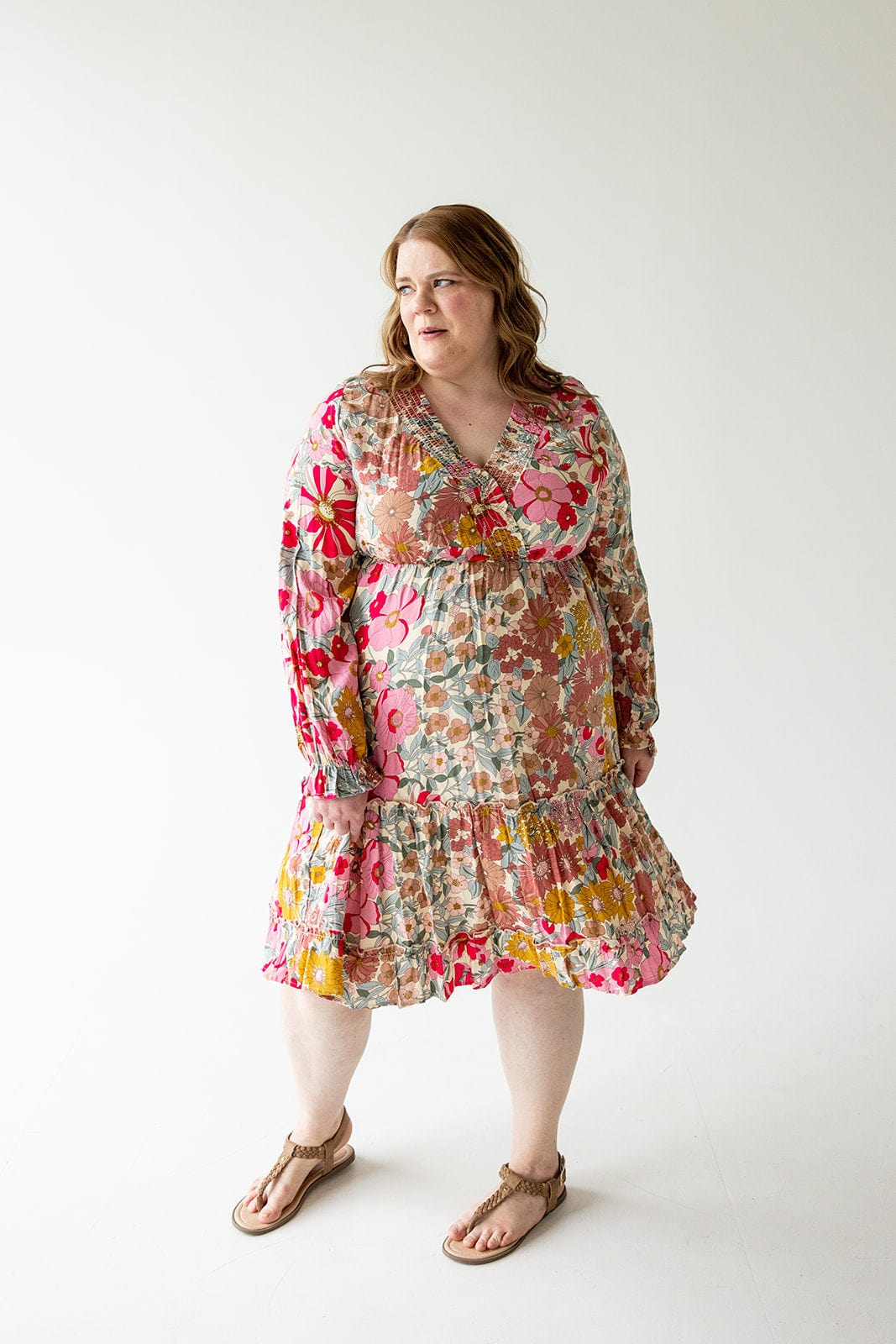 A person stands in Love Marlow's SMOCKED BODICE BOHO DRESS, showcasing pink, red, and yellow floral patterns, looking to the side.