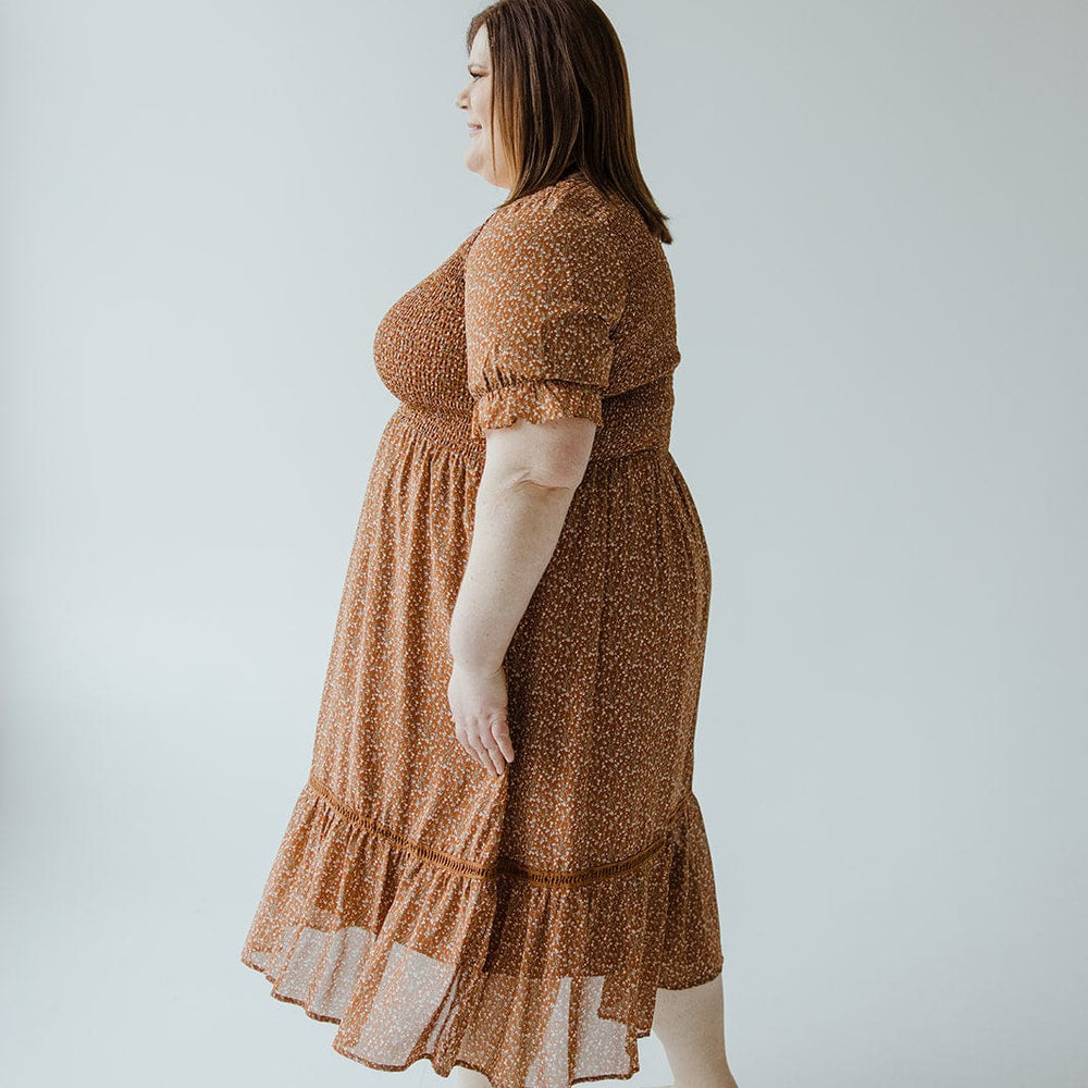 A person with straight brown hair is standing sideways in a studio, wearing the Mikarose SMOCKED BODICE DRESS WITH KICK HEM IN HARVEST PUMPKIN, paired with platform sandals. The background is plain and light-colored.