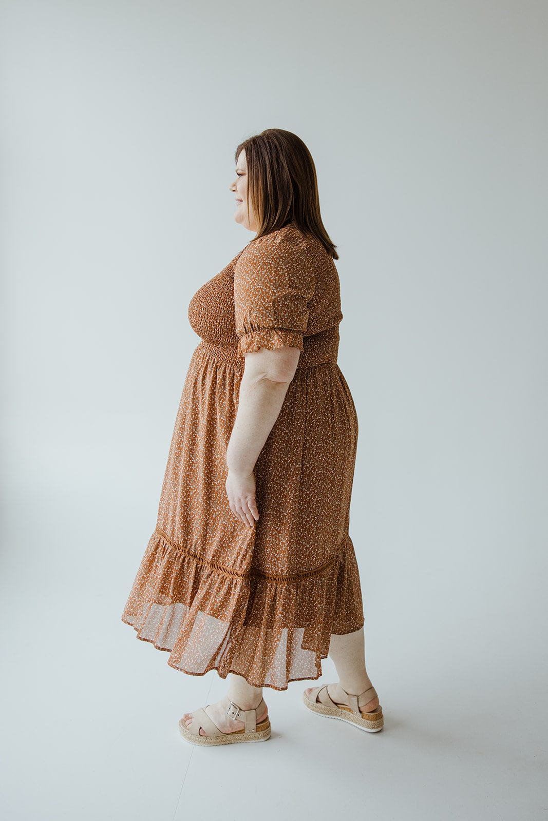 A person with straight brown hair is standing sideways in a studio, wearing the Mikarose SMOCKED BODICE DRESS WITH KICK HEM IN HARVEST PUMPKIN, paired with platform sandals. The background is plain and light-colored.