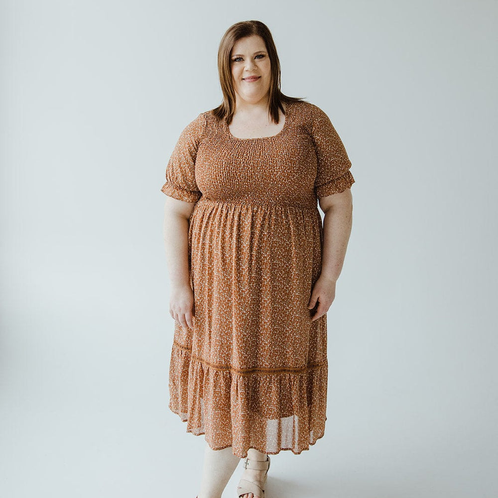 A woman in a "SMOCKED BODICE DRESS WITH KICK HEM IN HARVEST PUMPKIN" by Mikarose and sandals stands against a plain white background.