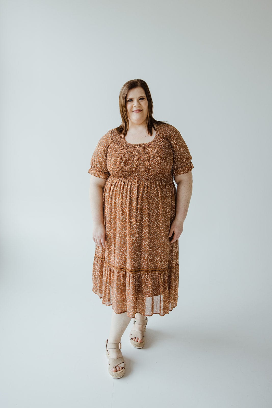 A woman in a "SMOCKED BODICE DRESS WITH KICK HEM IN HARVEST PUMPKIN" by Mikarose and sandals stands against a plain white background.