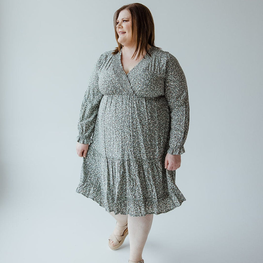 A woman stands against a plain background, wearing the SMOCKED BODICE MICRO FLORAL BOHO DRESS by Mikarose with beige sandals. She has shoulder-length hair and holds the hem of her dress with one hand.