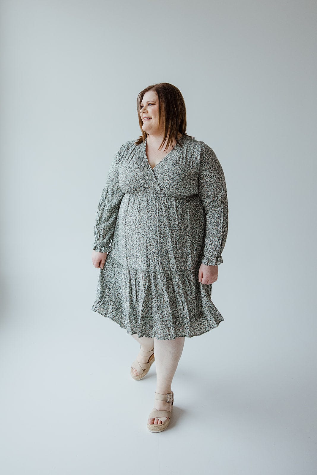 A woman stands against a plain background, wearing the SMOCKED BODICE MICRO FLORAL BOHO DRESS by Mikarose with beige sandals. She has shoulder-length hair and holds the hem of her dress with one hand.