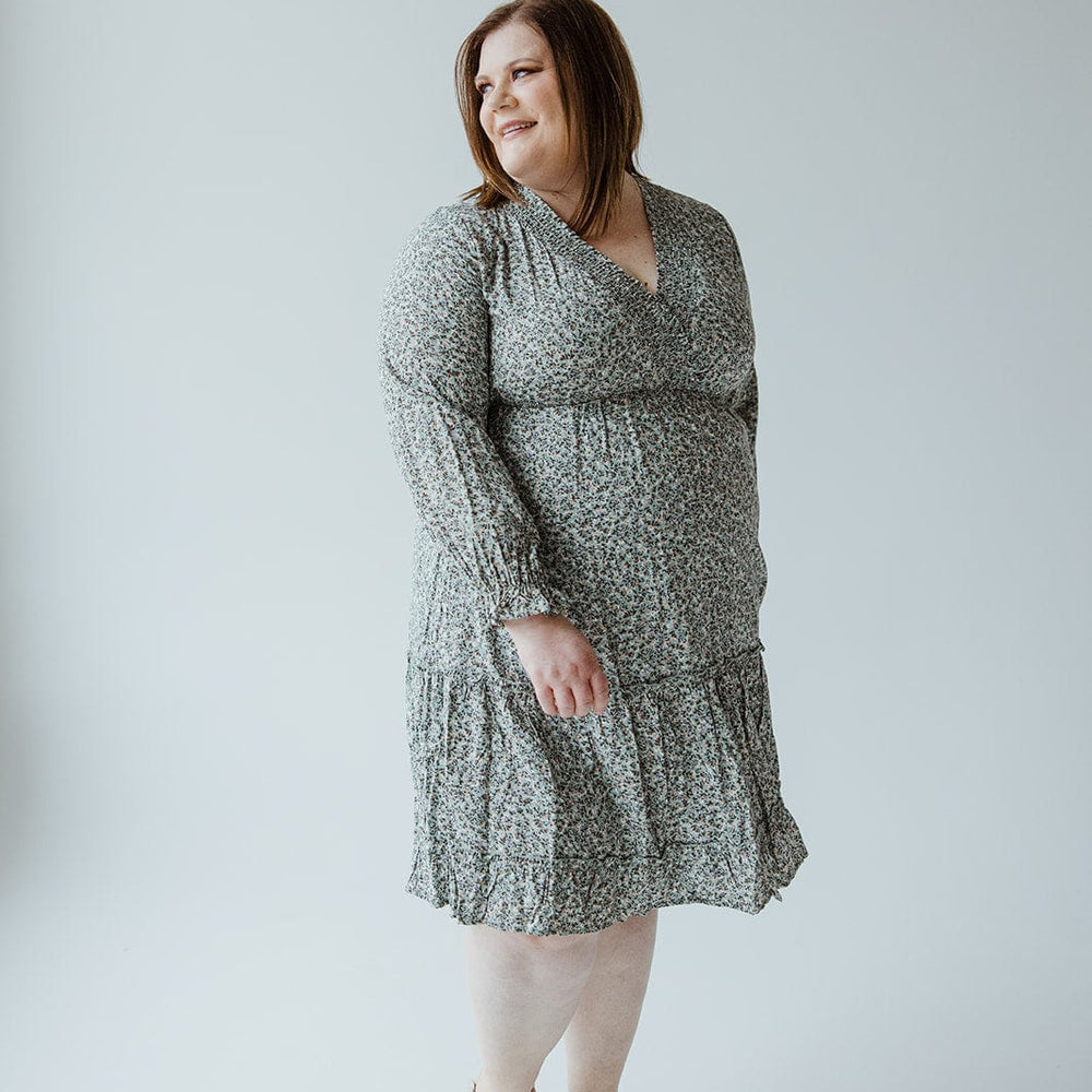 A woman stands, looking to the side, dressed in a grey-patterned Smocking Bodice Micro Floral Boho Dress by Mikarose, paired with light-colored sandals against a plain background.