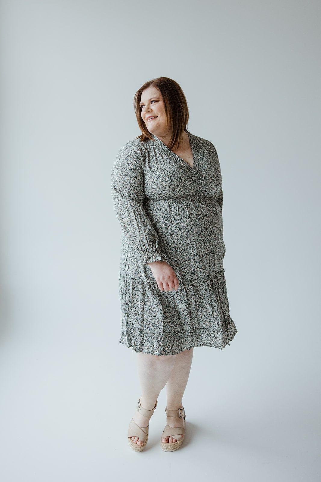 A woman stands, looking to the side, dressed in a grey-patterned Smocking Bodice Micro Floral Boho Dress by Mikarose, paired with light-colored sandals against a plain background.