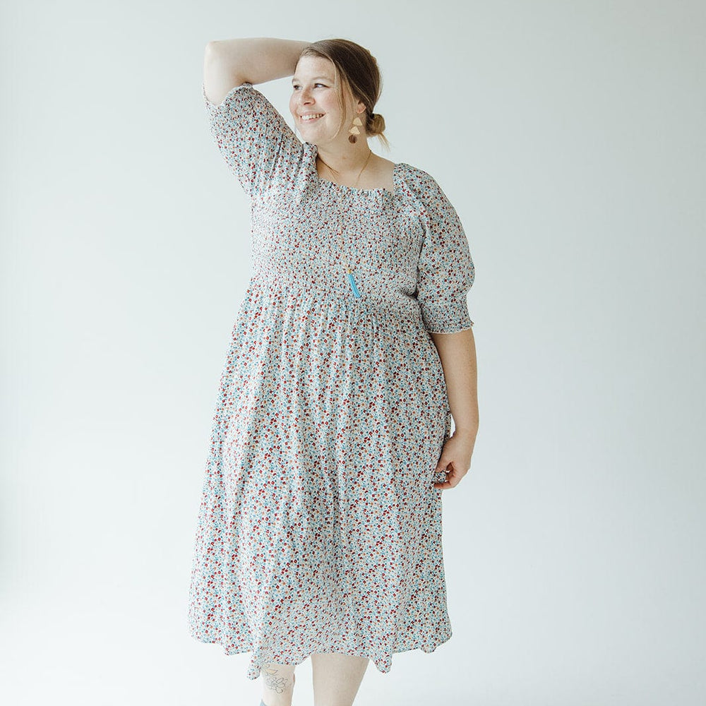 A woman in a Mikarose SMOCKED BODICE MIDI-LENGTH DRESS IN INDEPENDENCE with puff sleeves stands with one hand on her head, smiling. She is wearing light blue sandals and large earrings, with her hair tied back. The background is a plain white.