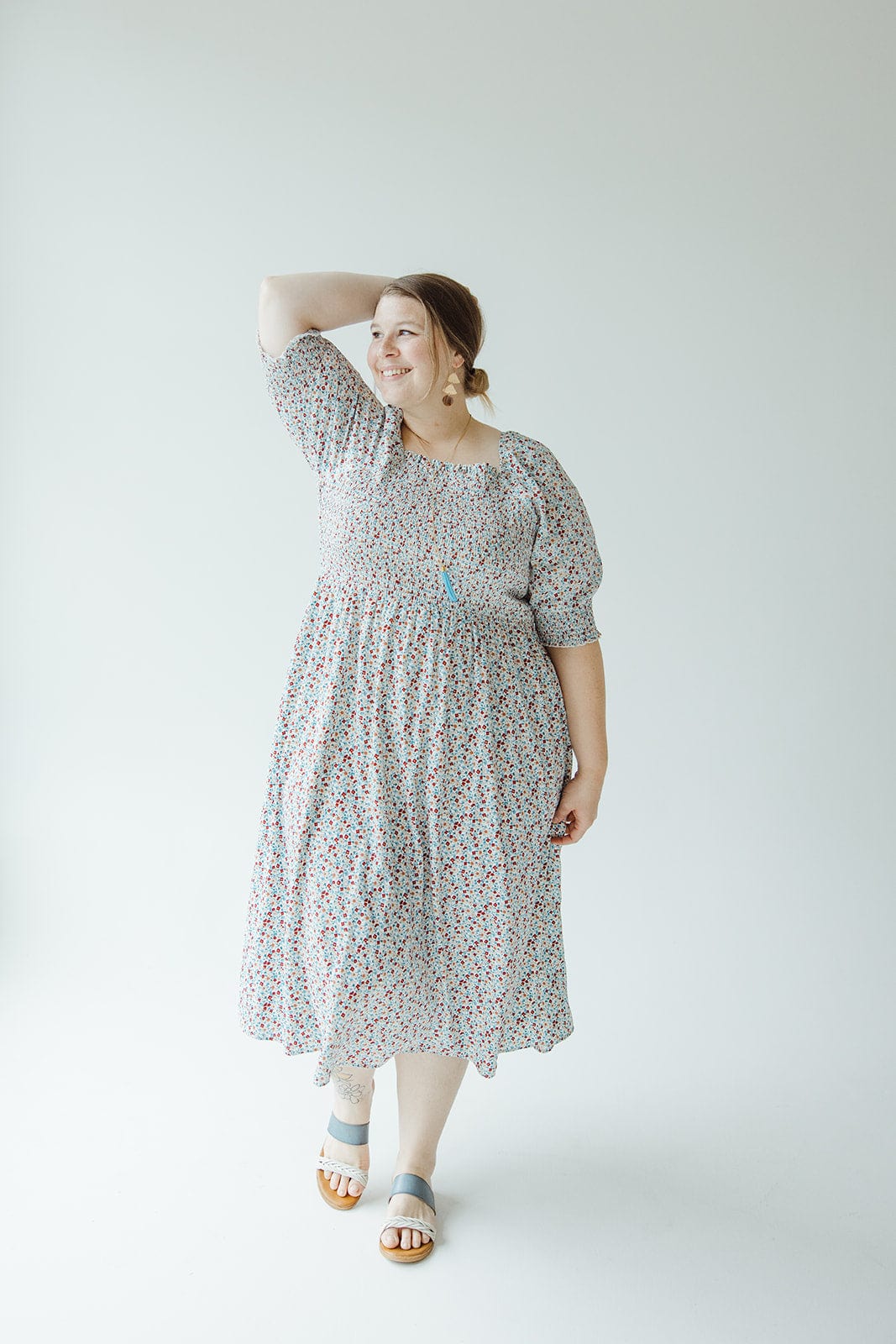 A woman in a Mikarose SMOCKED BODICE MIDI-LENGTH DRESS IN INDEPENDENCE with puff sleeves stands with one hand on her head, smiling. She is wearing light blue sandals and large earrings, with her hair tied back. The background is a plain white.