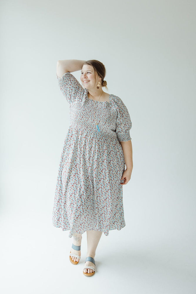 A woman in a Mikarose SMOCKED BODICE MIDI-LENGTH DRESS IN INDEPENDENCE with puff sleeves stands with one hand on her head, smiling. She is wearing light blue sandals and large earrings, with her hair tied back. The background is a plain white.