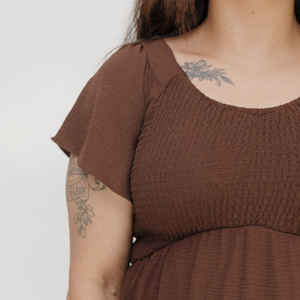 Person wearing a brown dress with tattoos on arms against a white background
