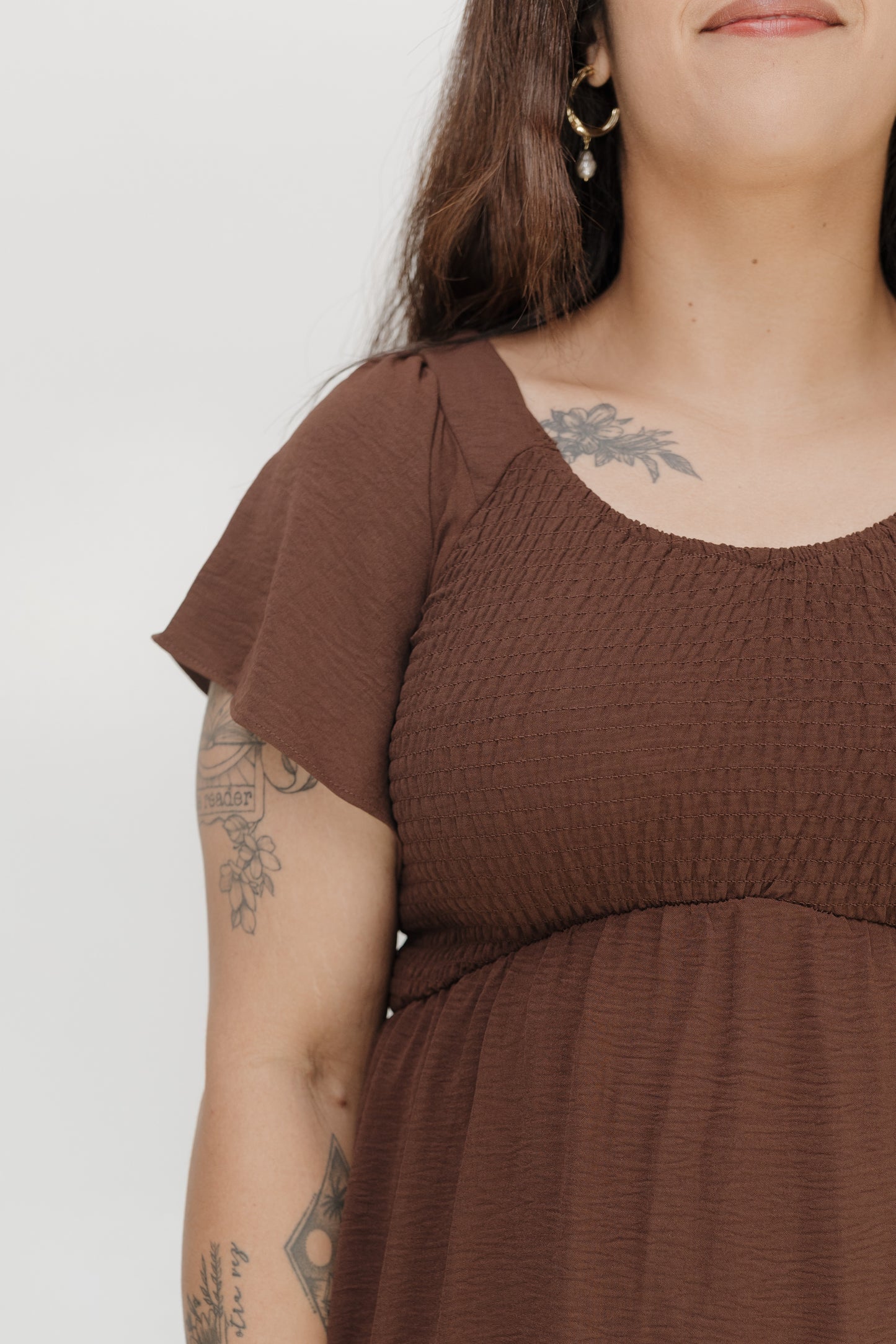 Person wearing a brown dress with tattoos on arms against a white background