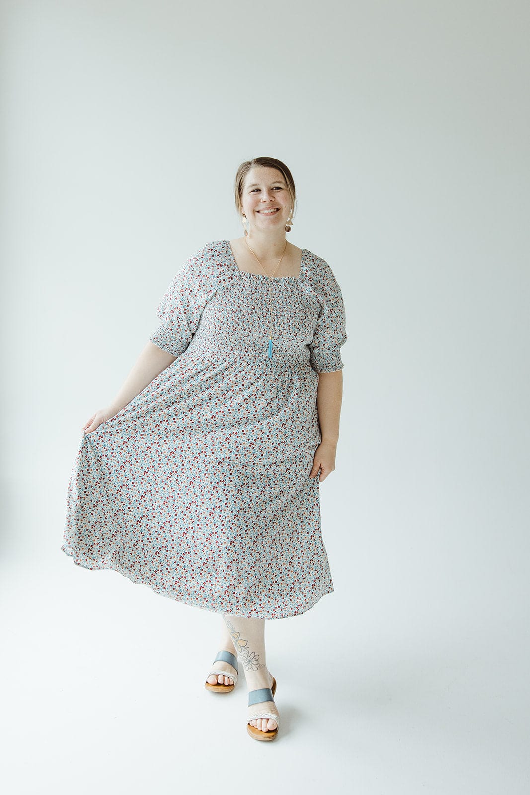 A woman stands in a SMOCKED BODICE MIDI-LENGTH DRESS IN INDEPENDENCE, smiling while holding the hem with one hand. She is wearing sandals and standing against a plain white background. The dress is from Mikarose.