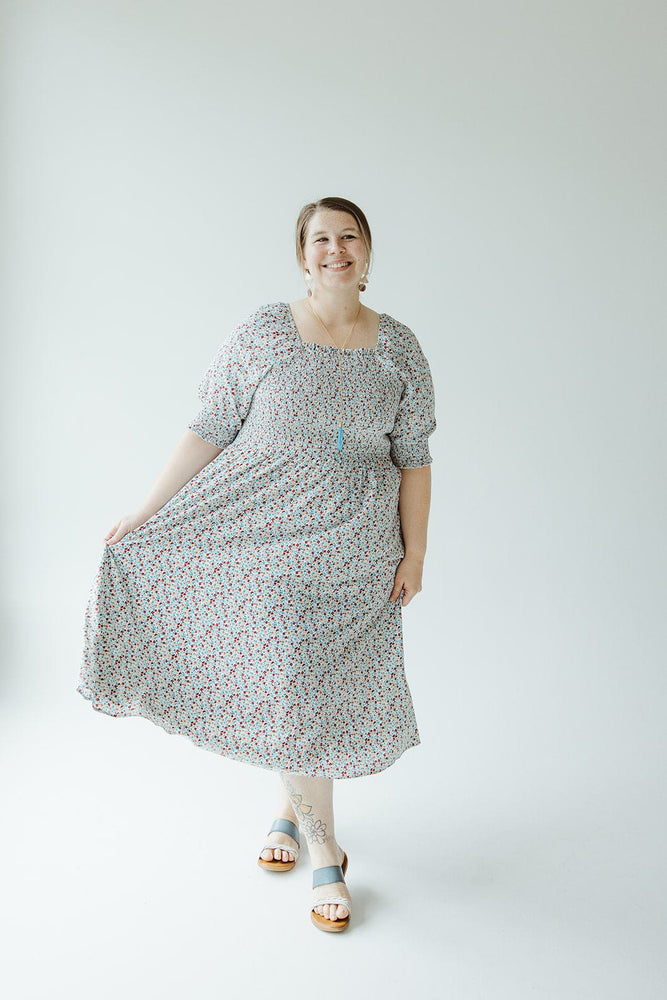 A woman stands in a SMOCKED BODICE MIDI-LENGTH DRESS IN INDEPENDENCE, smiling while holding the hem with one hand. She is wearing sandals and standing against a plain white background. The dress is from Mikarose.