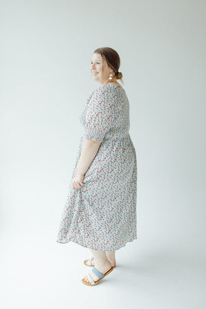 A woman in a Mikarose SMOCKED BODICE MIDI-LENGTH DRESS IN INDEPENDENCE with puff sleeves and sandals smiles, standing in a white, well-lit space.