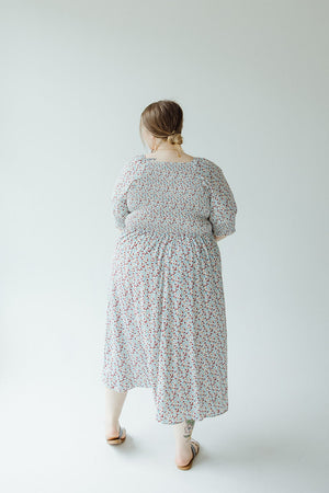 A person with light brown hair in a bun, wearing a Mikarose SMOCKED BODICE MIDI-LENGTH DRESS IN INDEPENDENCE and sandals, stands with their back facing the camera against a white background.