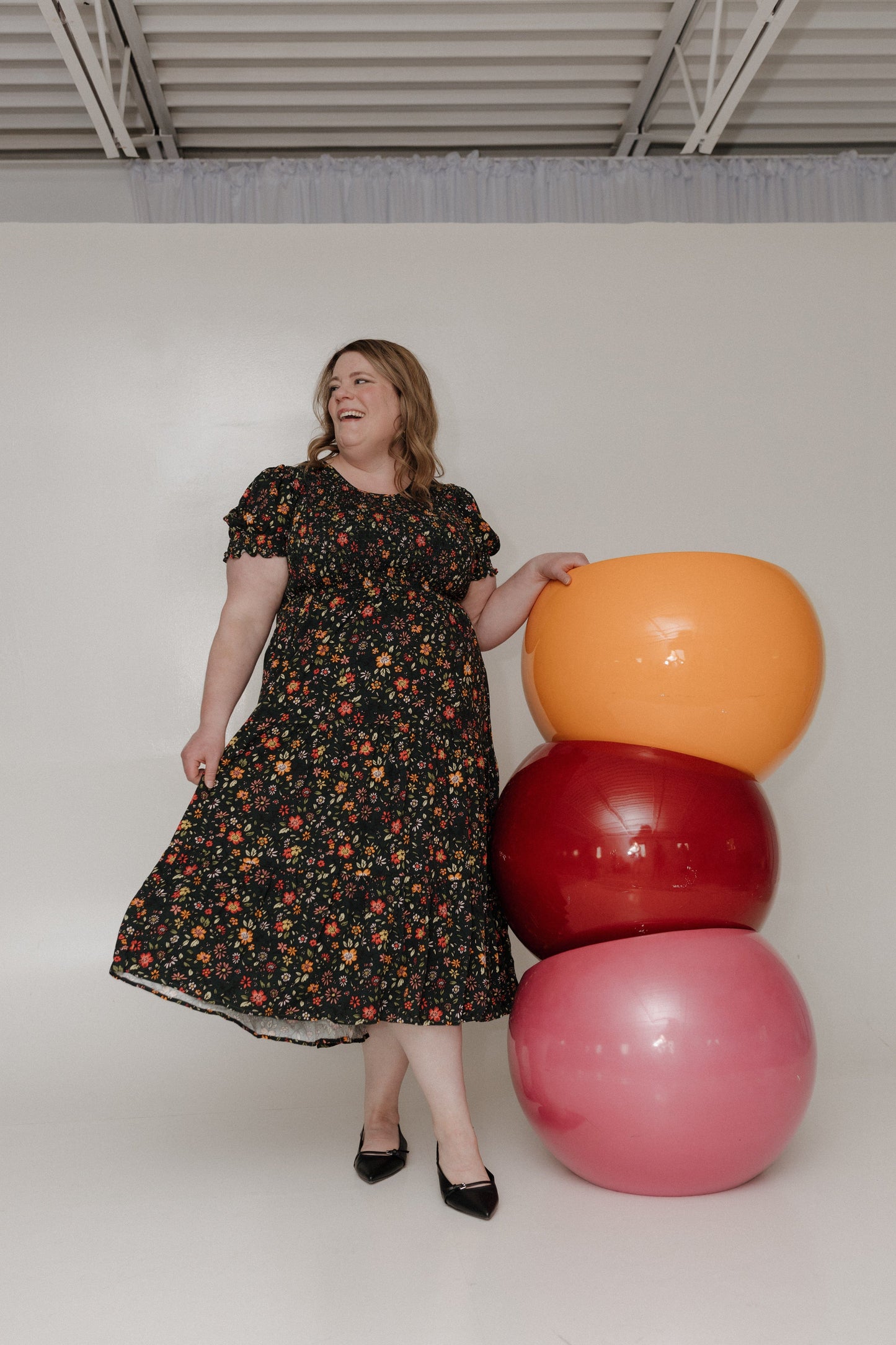 Woman in a floral dress standing next to stacked exercise balls in a minimal indoor setting.