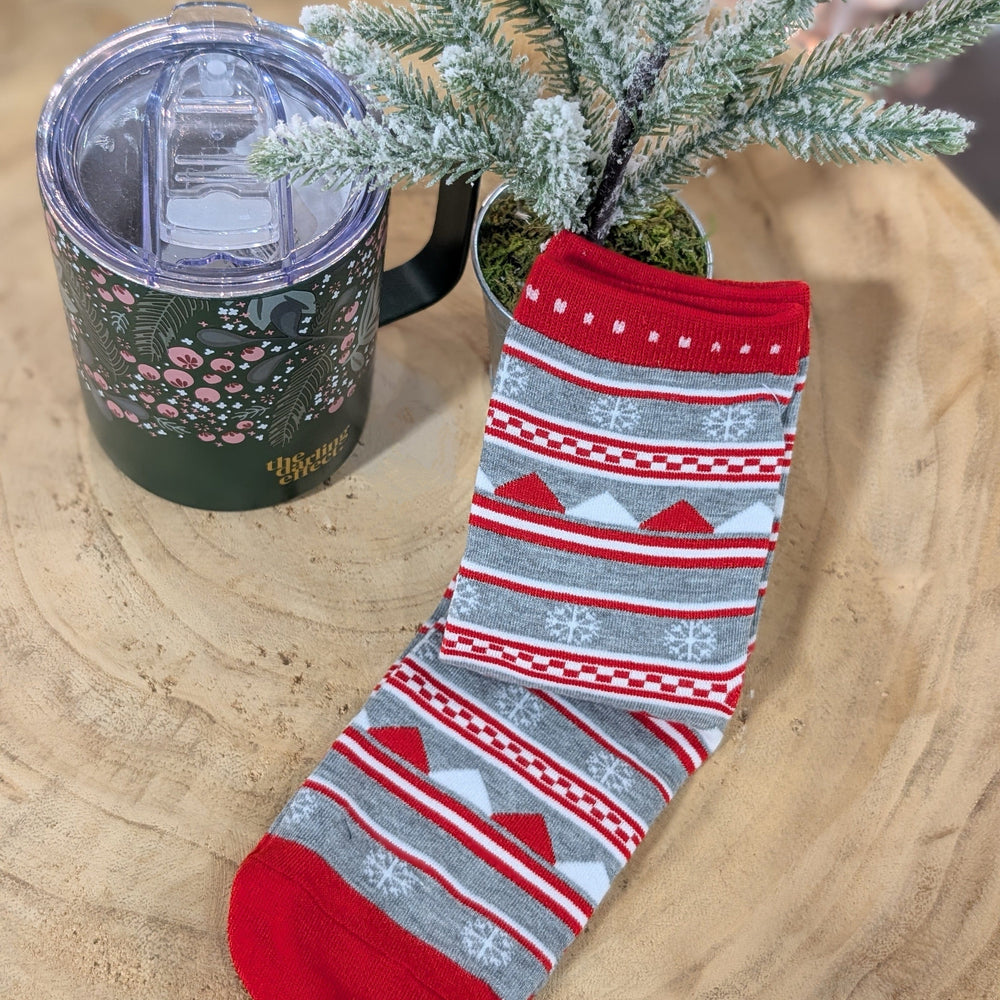 A pair of cozy festive socks, adorned with red, white, and gray snowflake patterns, rests warmly on a wooden surface beside a green mug with a lid and a small frosted artificial plant.
