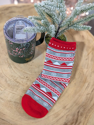 A pair of cozy festive socks, adorned with red, white, and gray snowflake patterns, rests warmly on a wooden surface beside a green mug with a lid and a small frosted artificial plant.