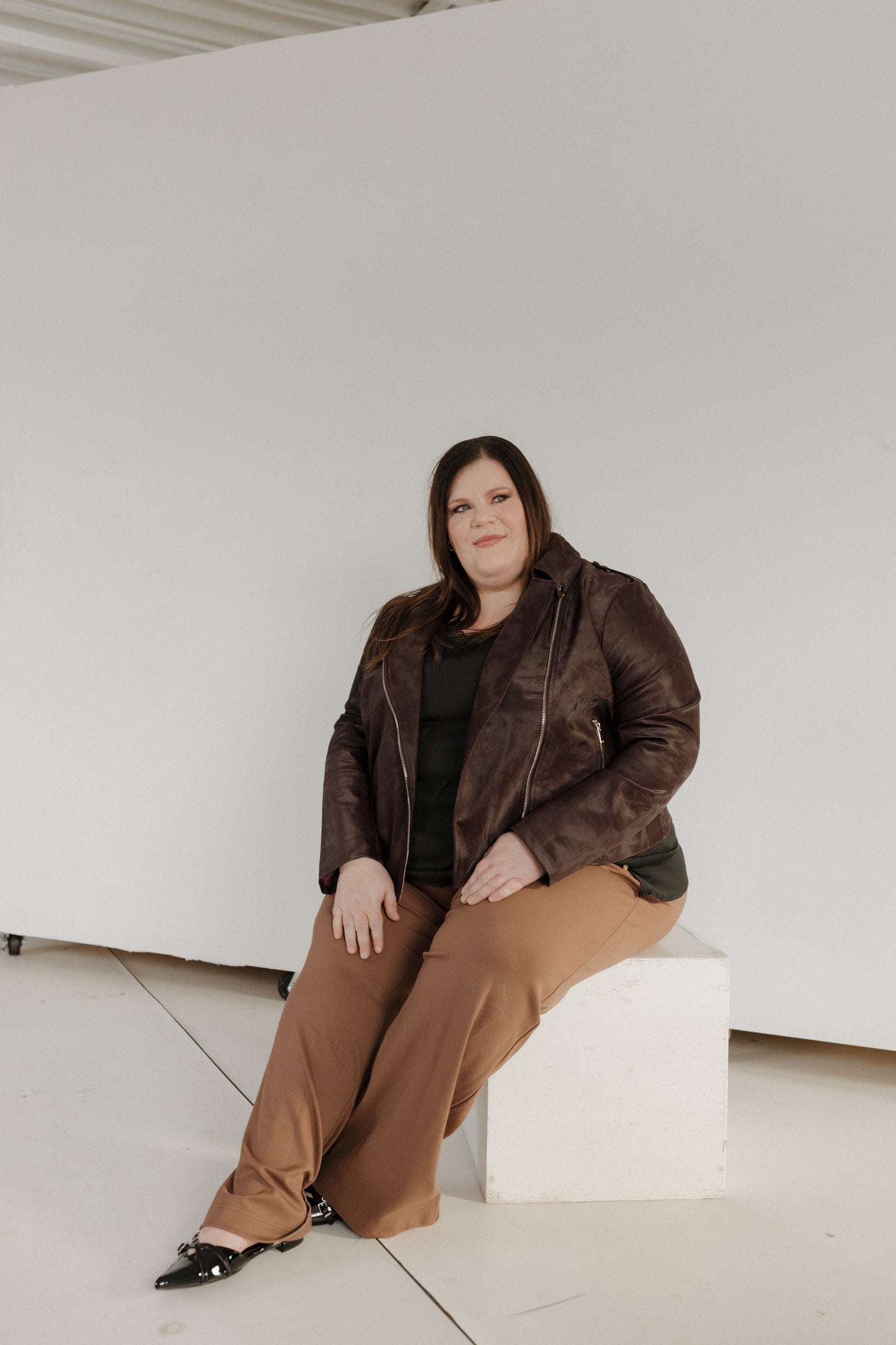 Woman in a brown leather jacket and pants sitting on a white block against a white wall.