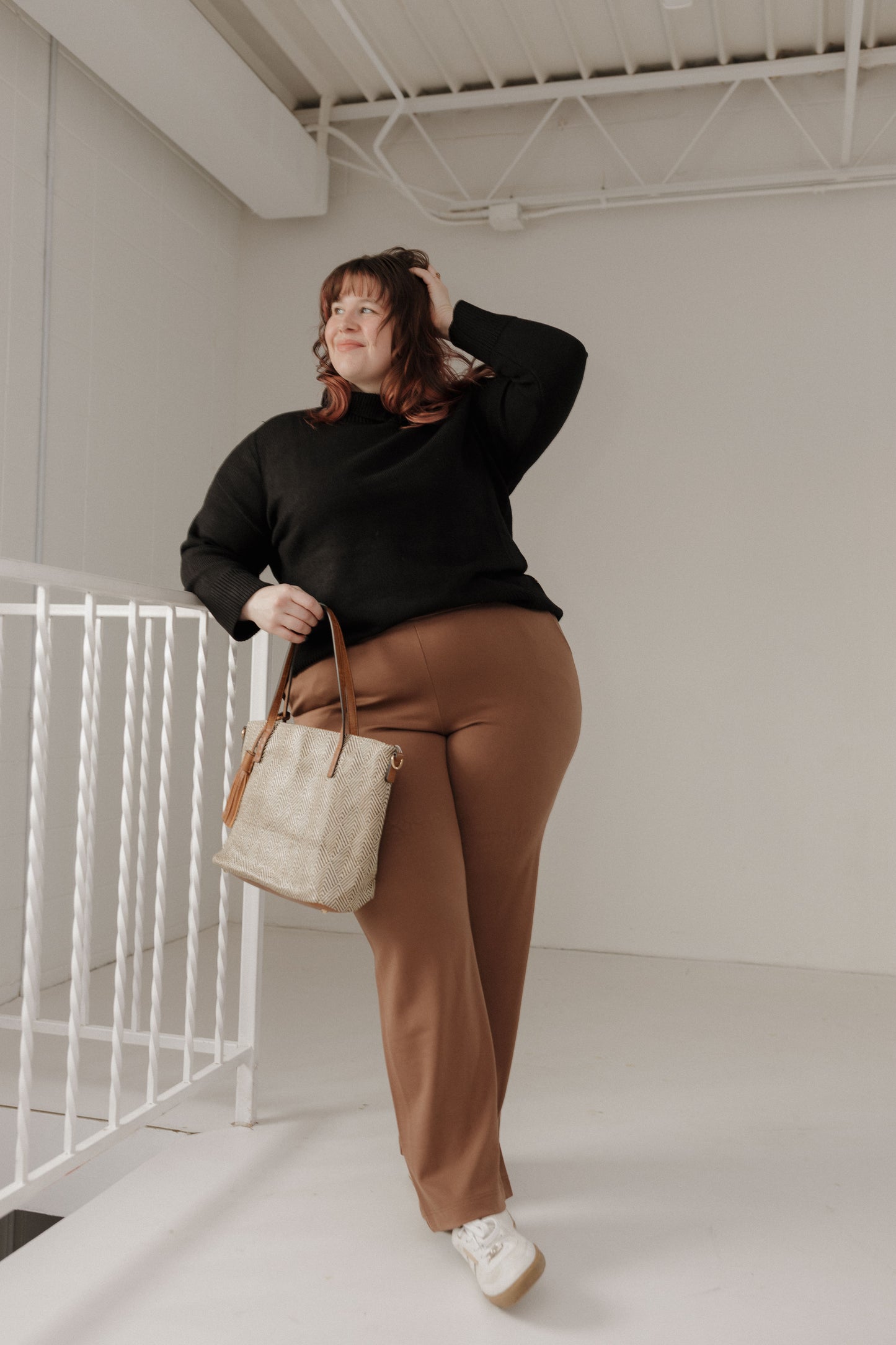 Person wearing a black sweater and brown pants holding a beige bag in a minimalistic setting.