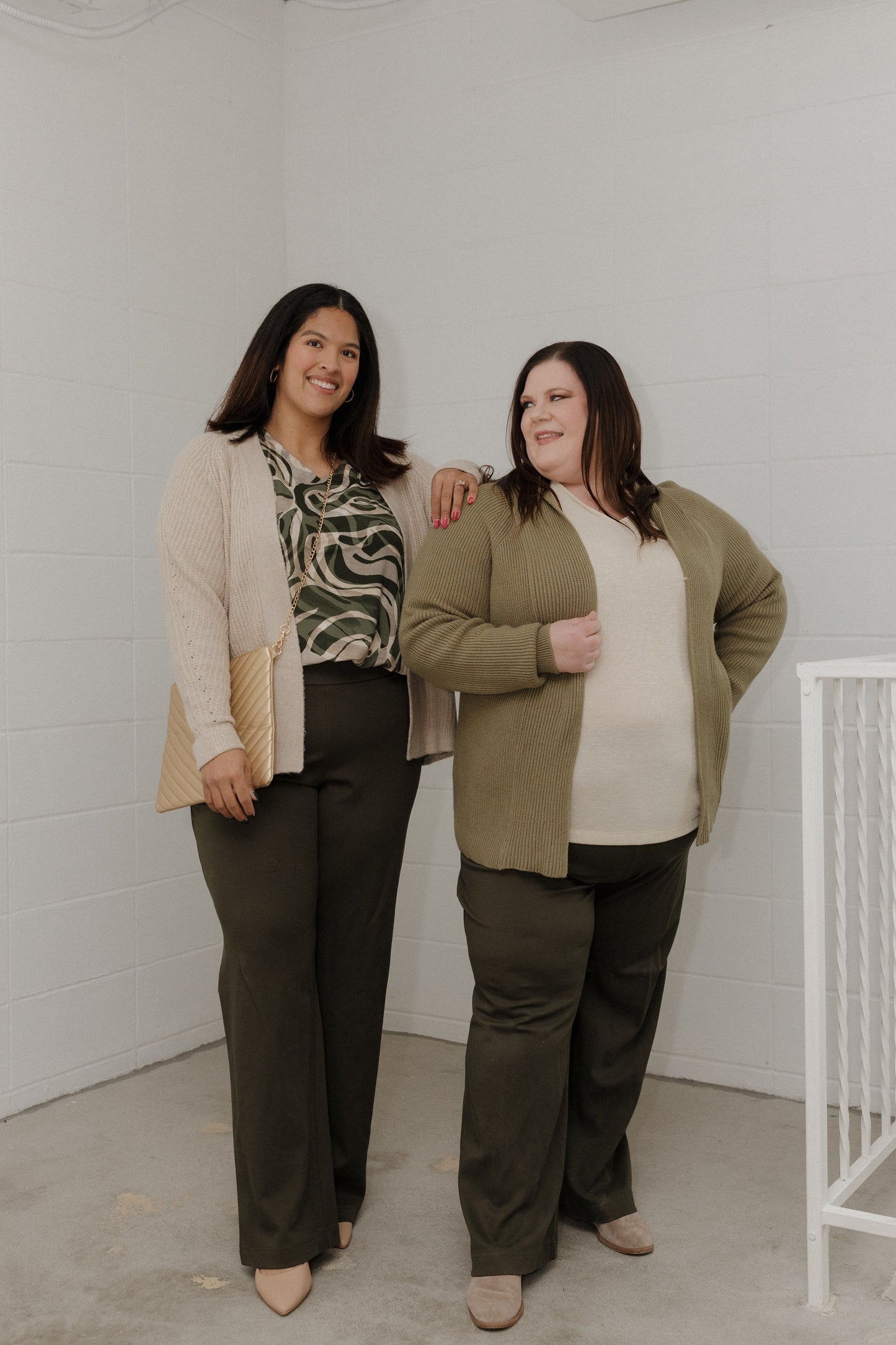 Two women standing together in a room with a white wall and a white crib.