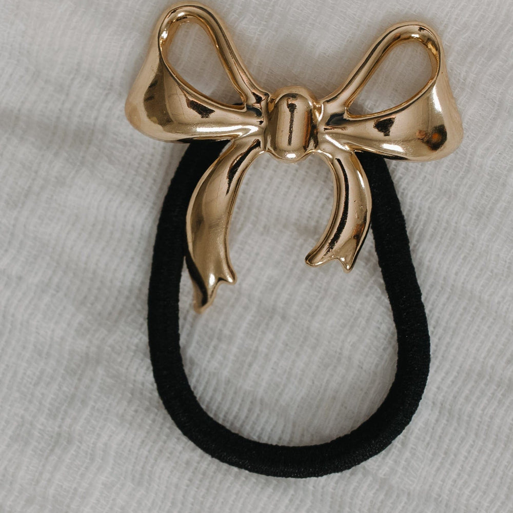 Gold bow-shaped hair accessory with a black rubber band on a gray fabric background