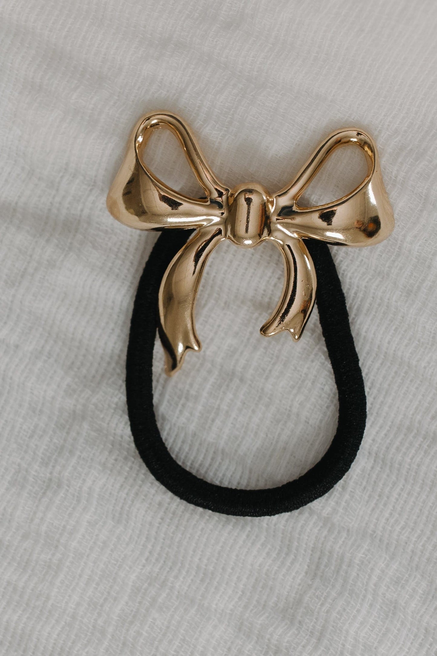 Gold bow-shaped hair accessory with a black rubber band on a gray fabric background