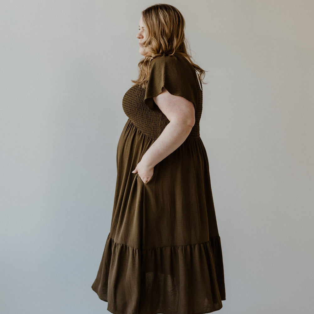 A person in a Bronzed Laurel midi dress, featuring a smocked bodice and flutter sleeves, with black slip-on shoes stands against a plain background.