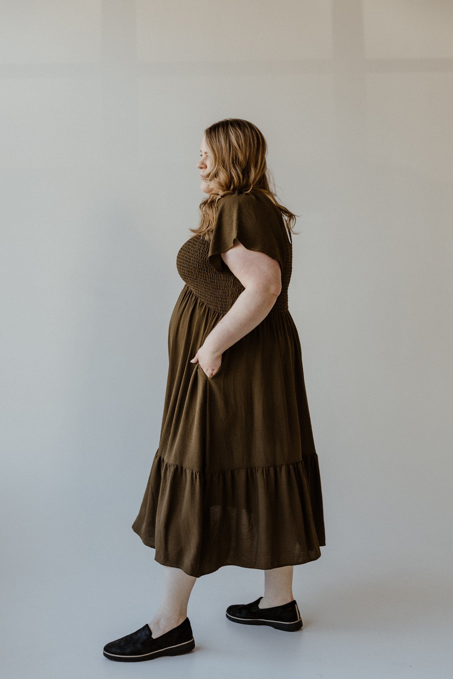 A person in a Bronzed Laurel midi dress, featuring a smocked bodice and flutter sleeves, with black slip-on shoes stands against a plain background.