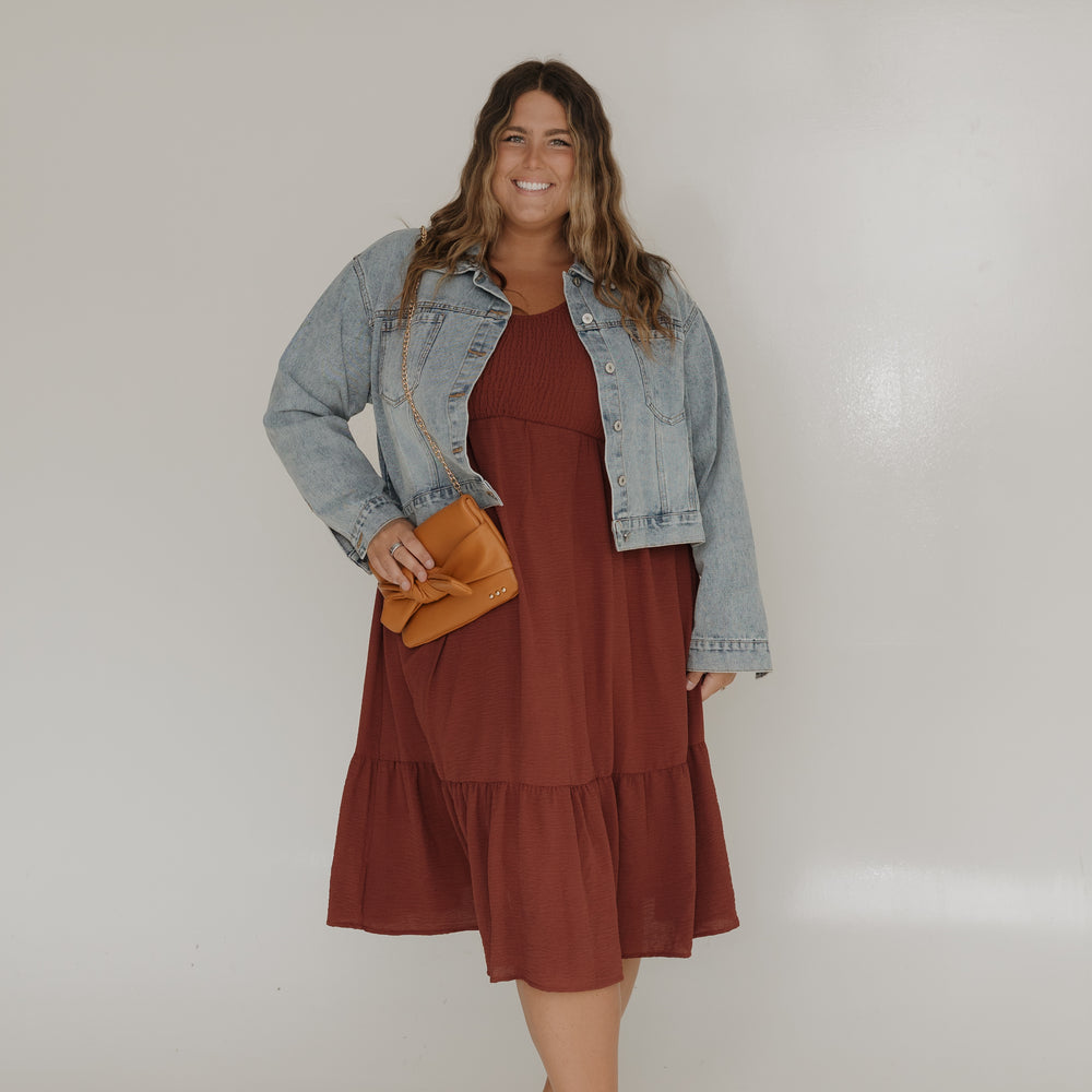 Woman wearing a brown dress with a denim jacket and holding a yellow clutch against a white background