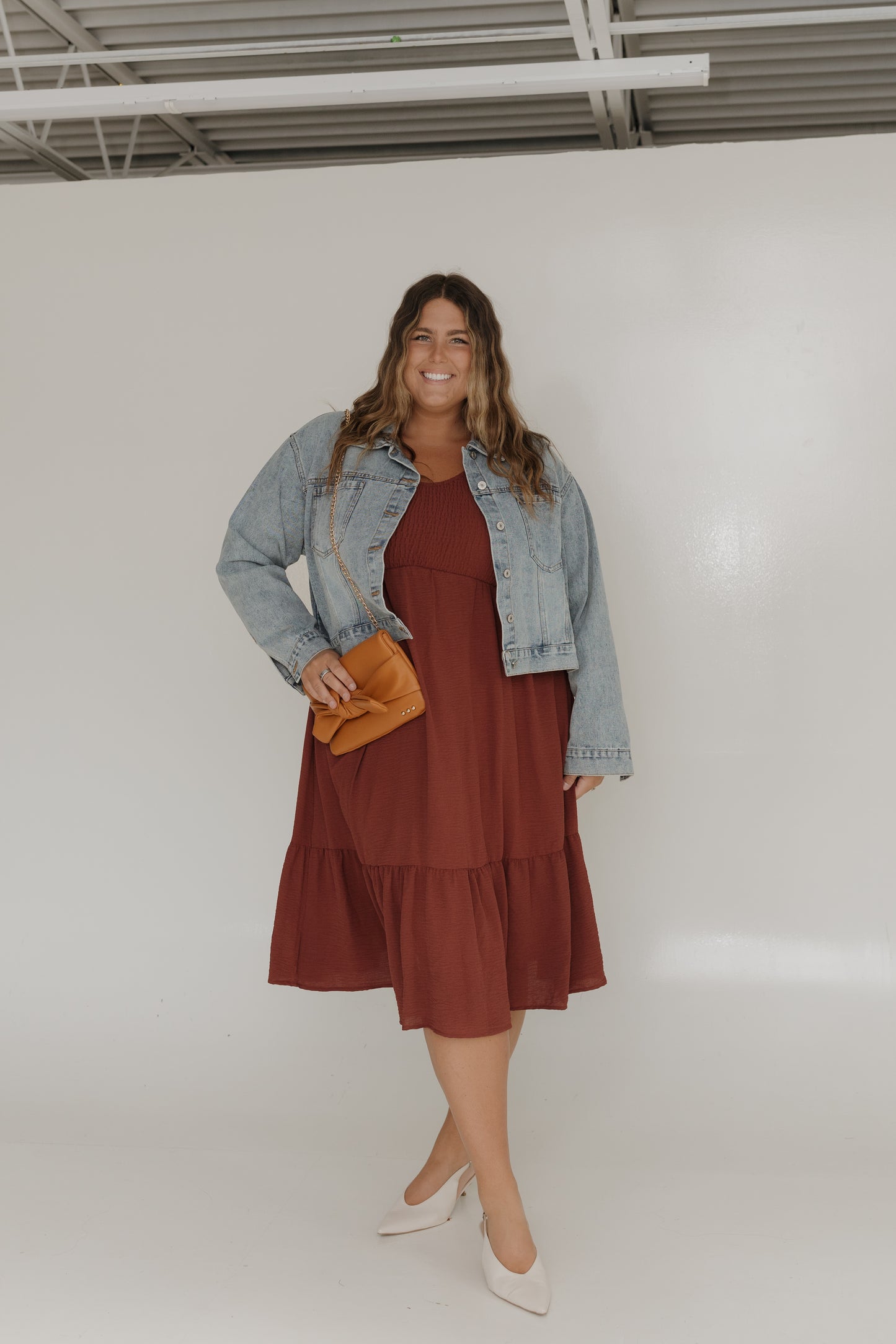 Woman wearing a brown dress with a denim jacket and holding a yellow clutch against a white background