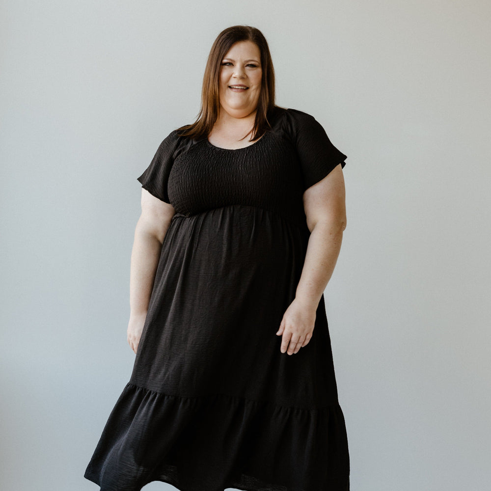 A person wearing a long black midi dress with flutter sleeves and matching black shoes stands against a plain light background, smiling.