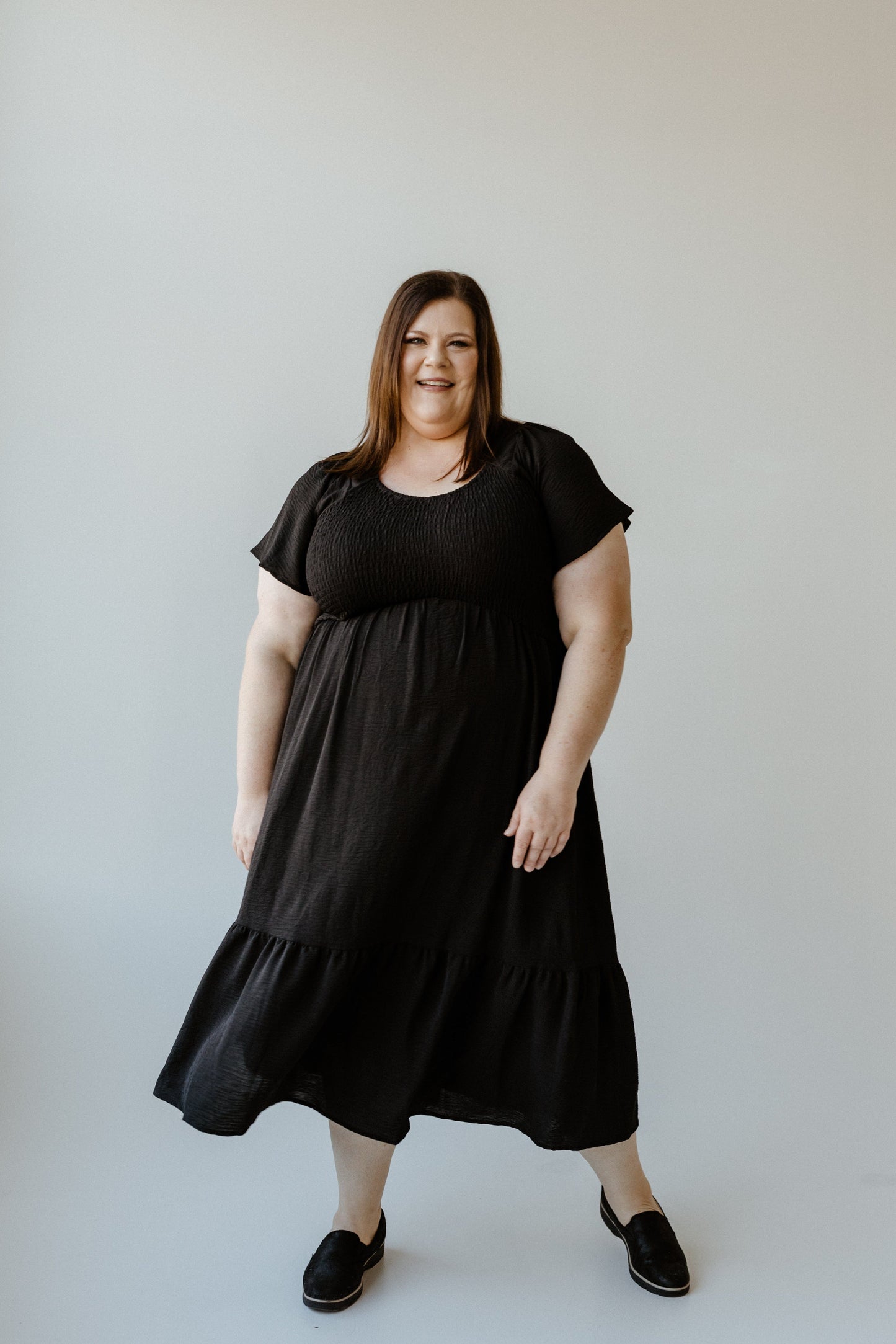 A person wearing a long black midi dress with flutter sleeves and matching black shoes stands against a plain light background, smiling.