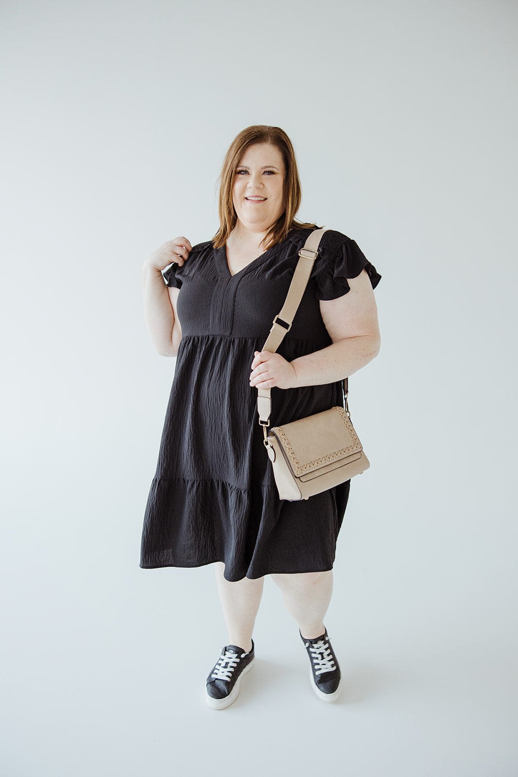 A woman is standing in the SOLID WOVEN TIERED DRESS WITH FLUTTER SLEEVE IN BLACK by Oddi, paired with black and white sneakers and a beige crossbody bag, against a plain white background. She is smiling and holding the bag strap with one hand.