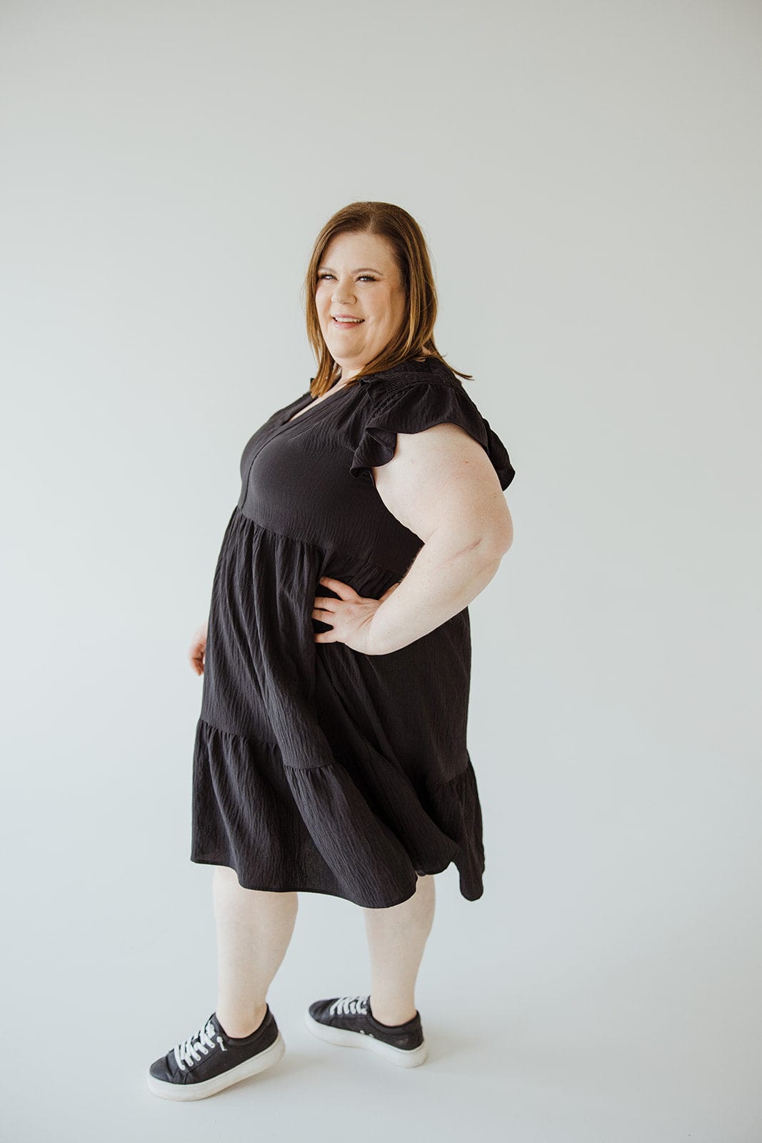 A person stands confidently wearing an Oddi Solid Woven Tiered Dress with Flutter Sleeves in Black and black sneakers against a plain white background.