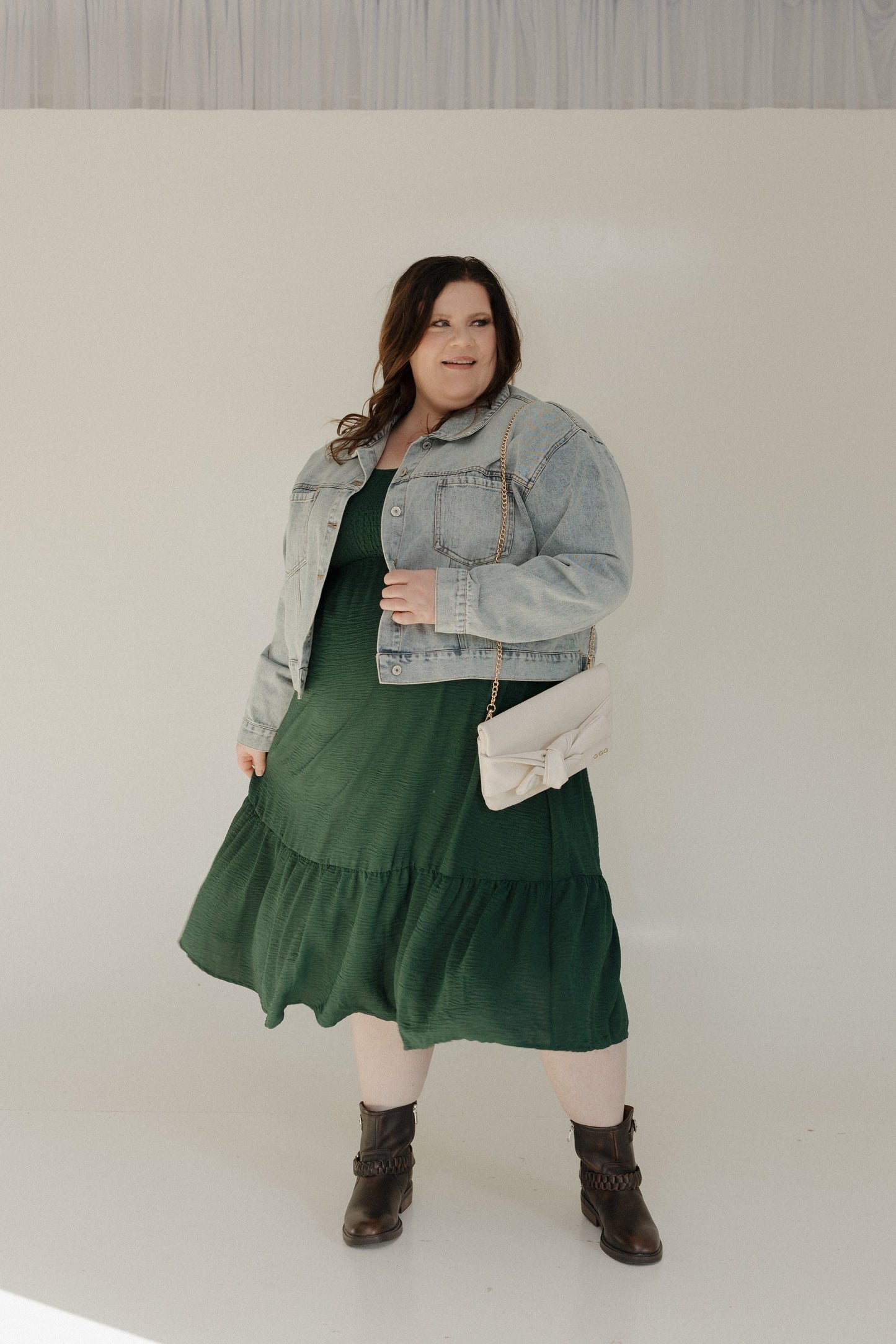 Woman wearing a green dress, denim jacket, and brown boots against a plain background