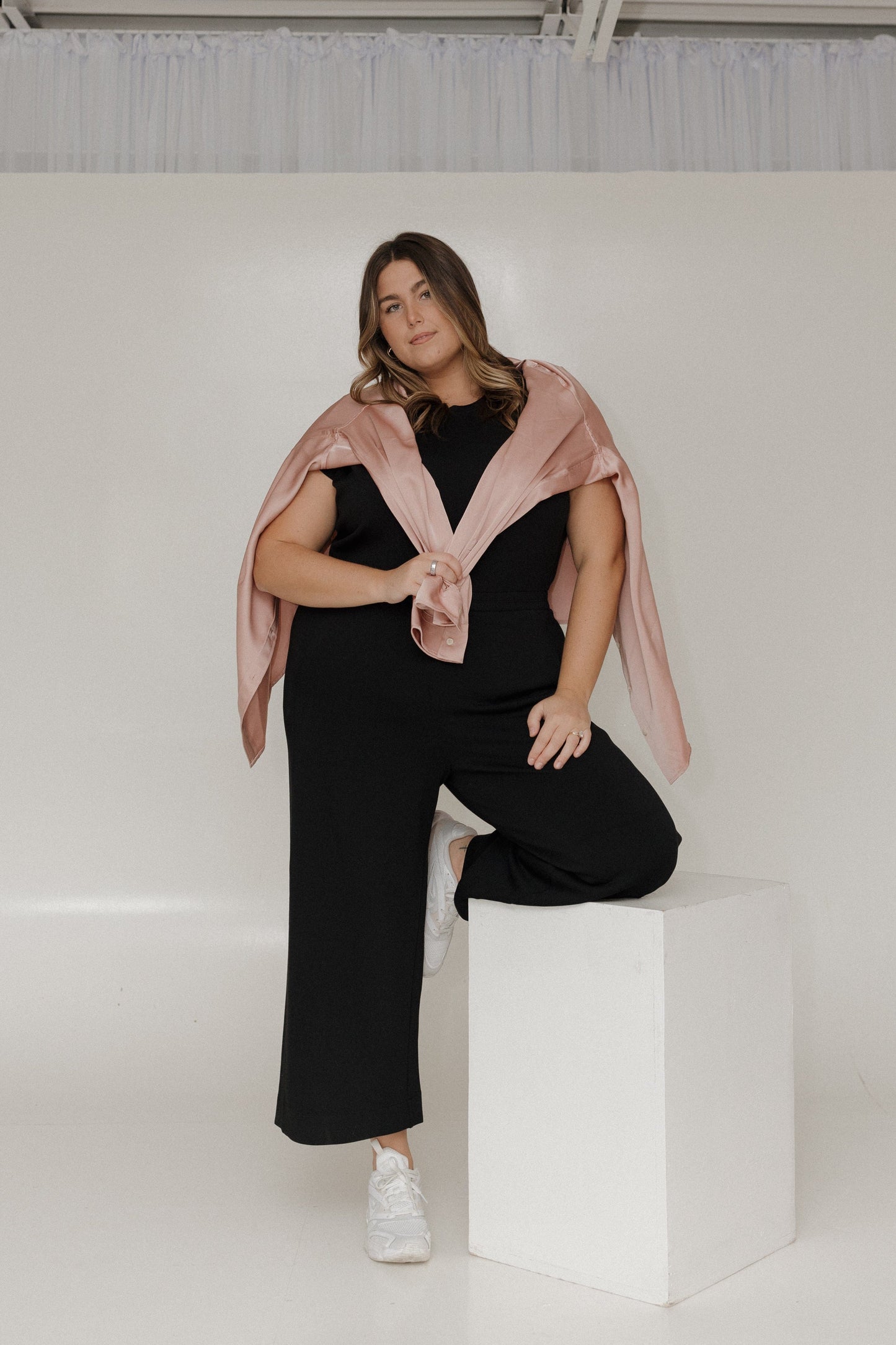 Woman wearing a black outfit with a pink scarf sitting on a white cube against a white background