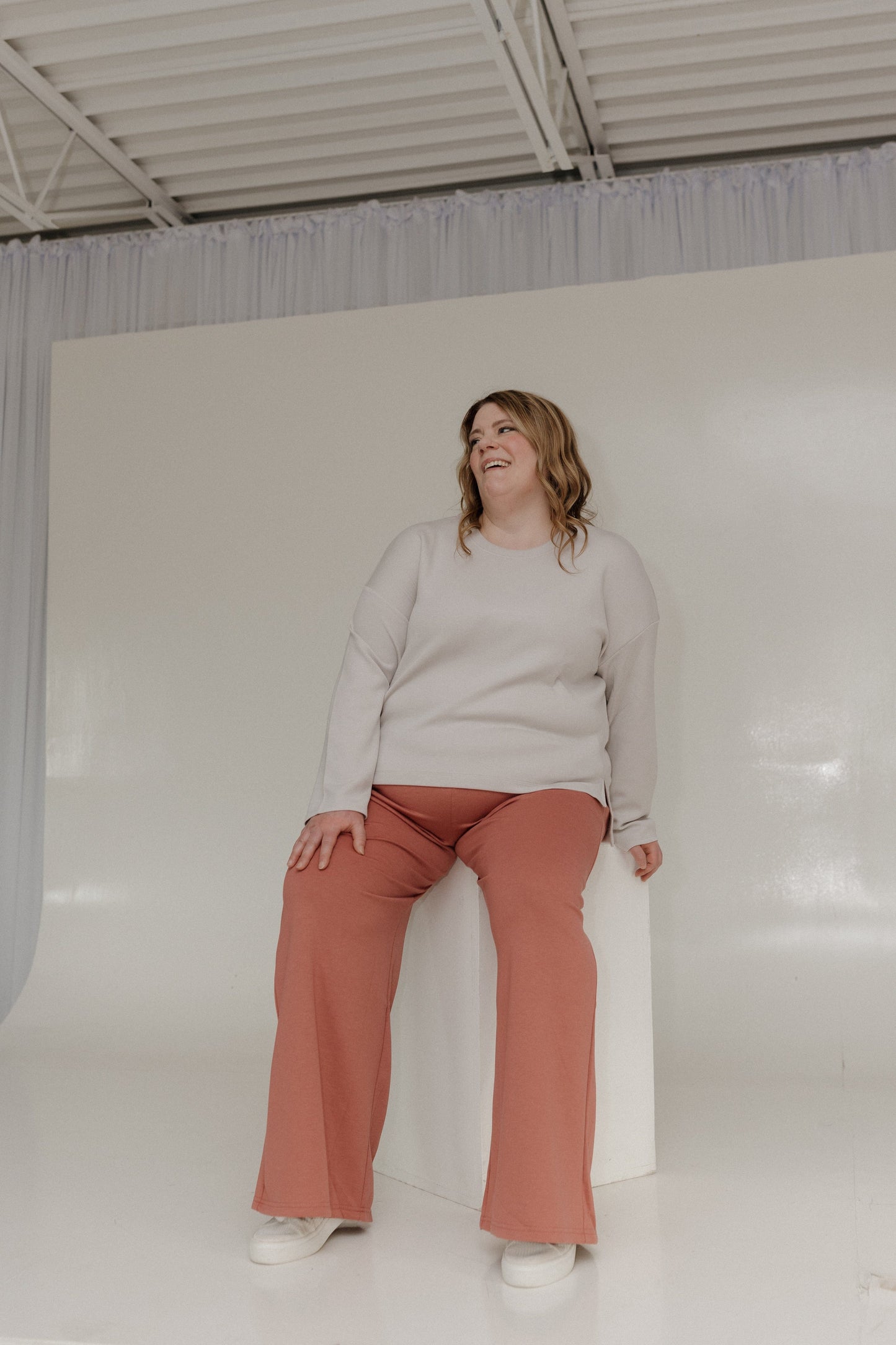 Woman wearing a light gray sweater and pink pants sitting on a white block against a white background