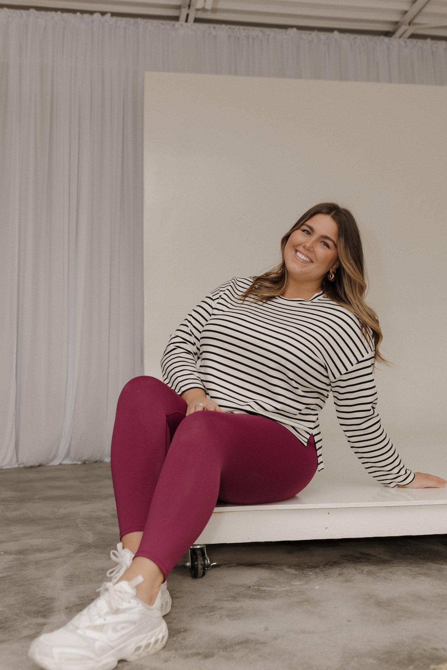 Woman sitting on a white bench wearing a striped sweater and maroon leggings.