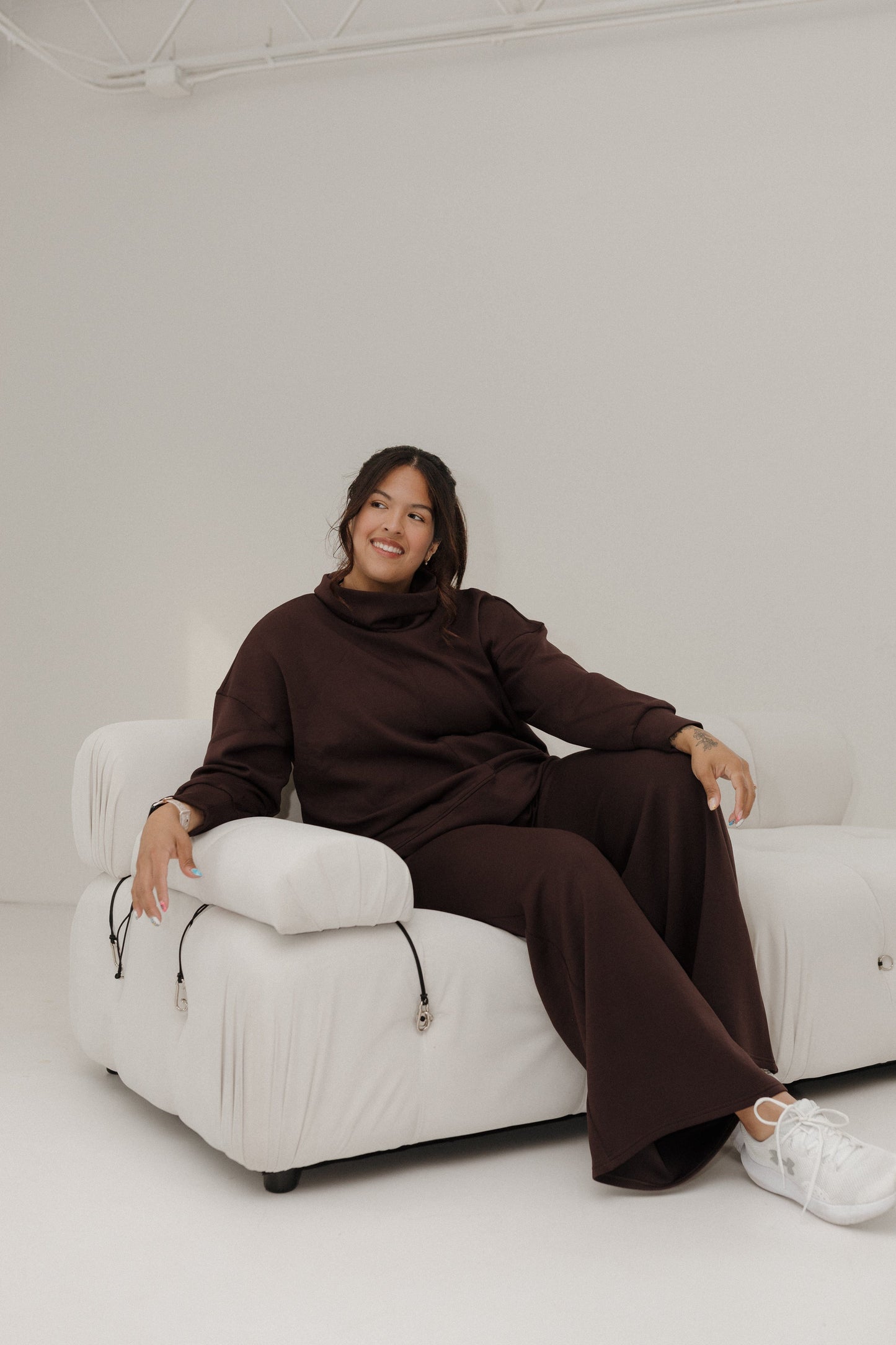 Person sitting on a white chair wearing a brown outfit in a minimalistic setting