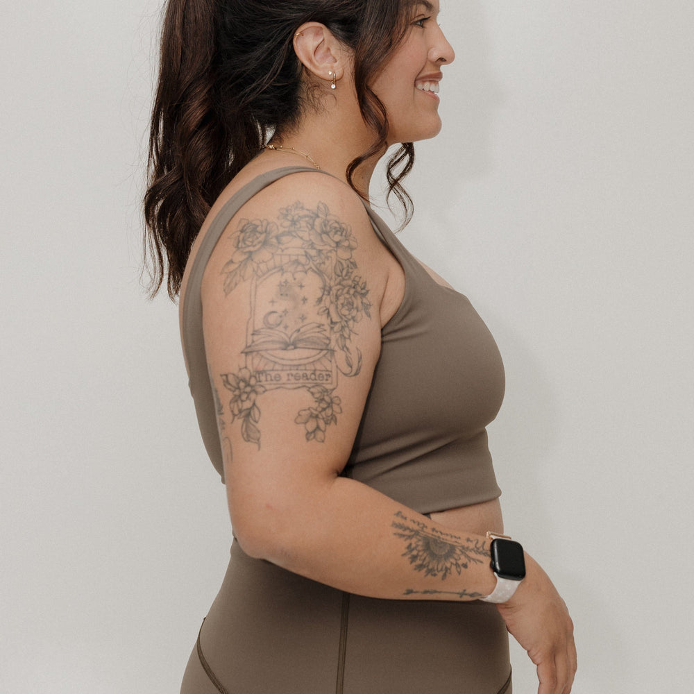 Woman wearing a brown athletic outfit with tattoos on her arms against a plain background