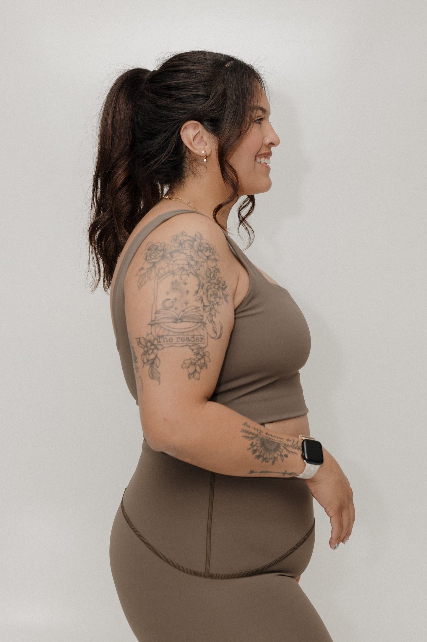 Woman wearing a brown athletic outfit with tattoos on her arms against a plain background