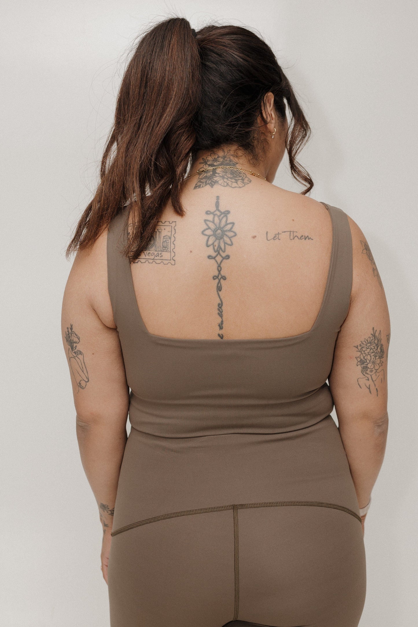 Person wearing a brown tank top and pants with visible tattoos on a plain background