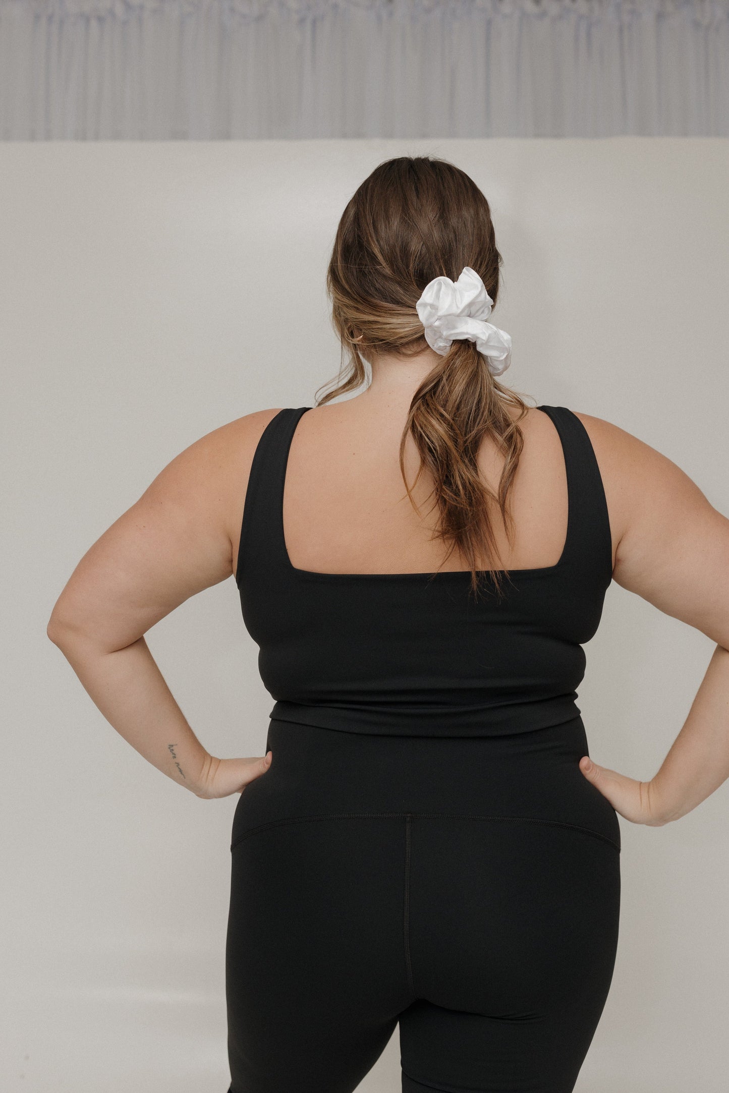 Person wearing a black tank top and leggings with a white scrunchie in their hair against a plain background