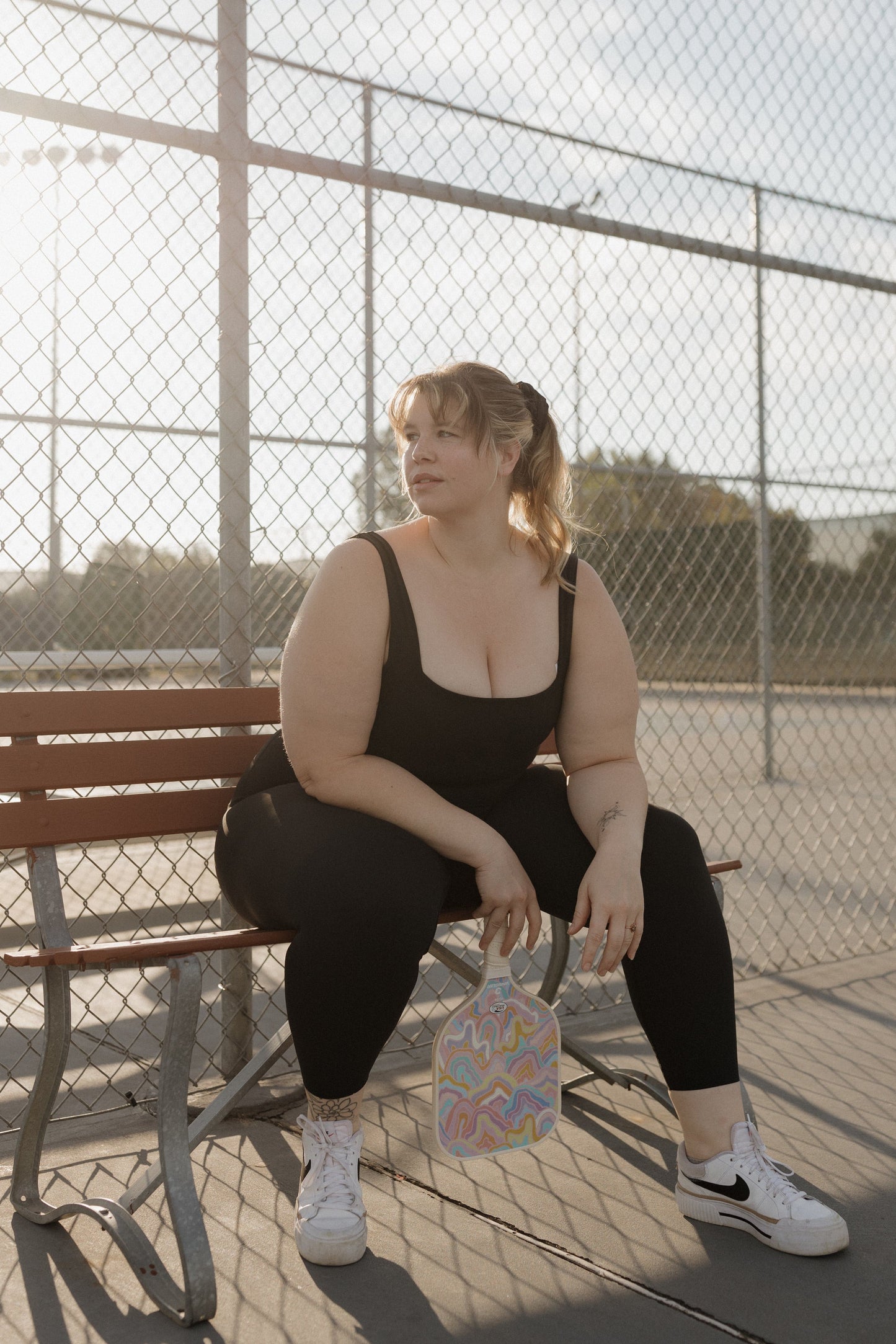 Woman sitting on a bench with a colorful bag, wearing a black outfit, in an outdoor setting.