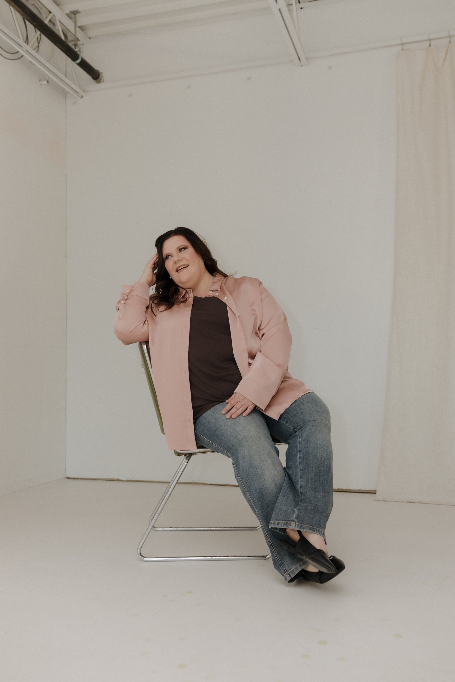 Woman sitting on a chair wearing a pink coat and jeans in a minimalistic room.