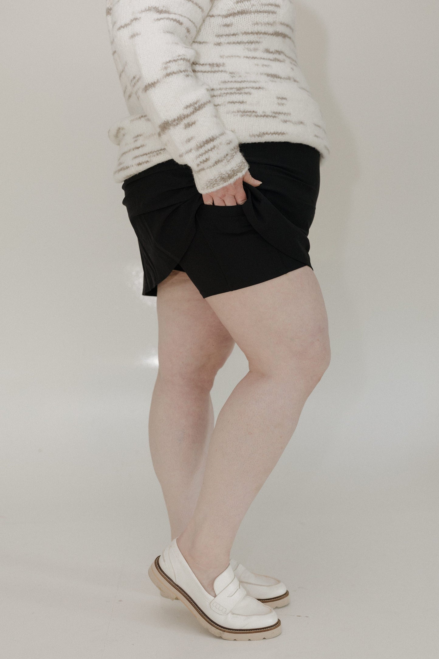 Person wearing a white sweater, black shorts, and white shoes on a plain background