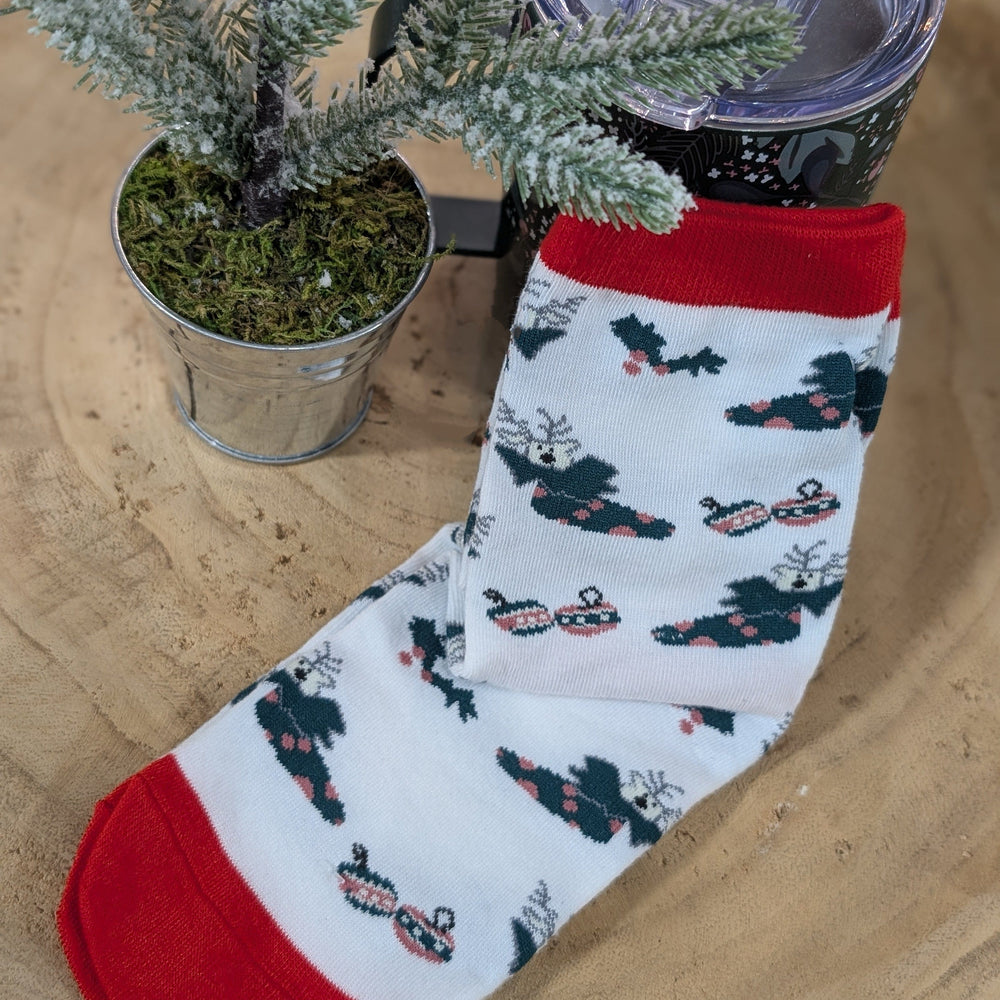 A cozy footwear piece, this holiday sock features a festive reindeer and sleigh design. It's perfectly displayed on a wooden surface, accompanied by a small potted Christmas tree and a tumbler.